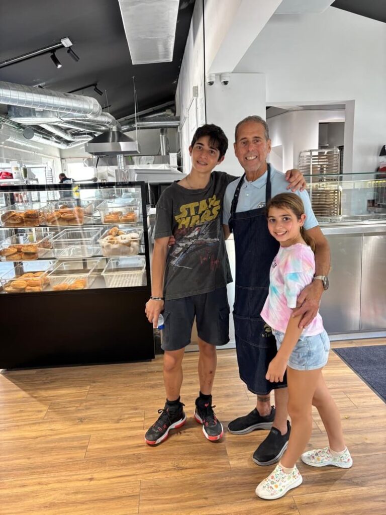 A man in an apron stands inside a bakery with a teenage boy and a young girl, with pastries and authentic New York bagels in Spain displayed in glass cases behind them.