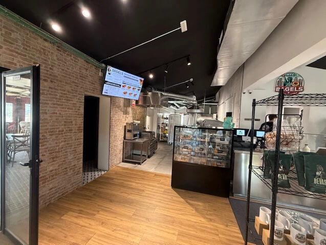 Interior of a bagel shop with a glass display case, merchandise shelf, exposed brick walls, wood flooring, and a digital menu overhead.